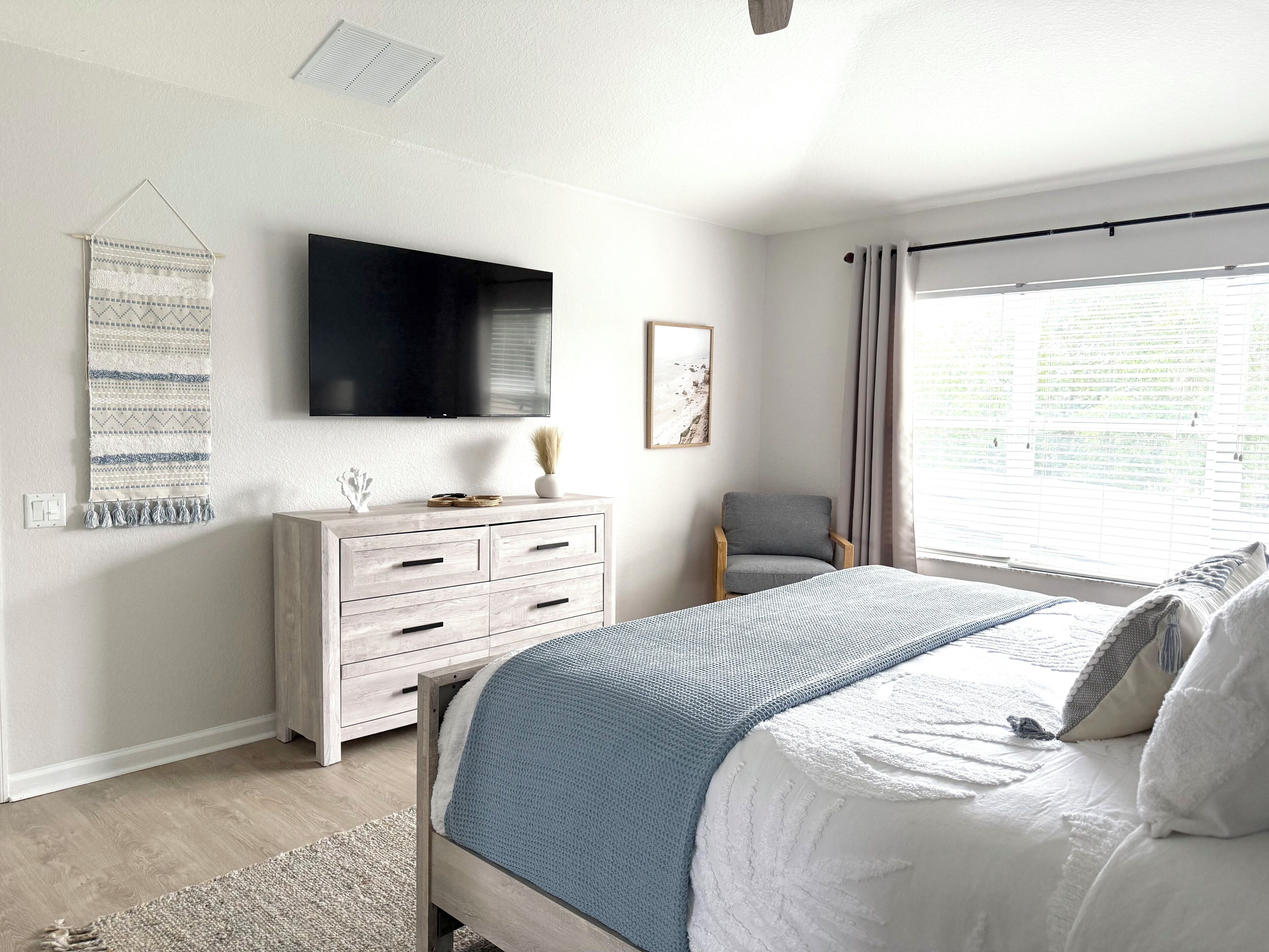 Une chambre à coucher avec un lit, une commode, une chaise, un téléviseur et une fenêtre avec des rideaux.