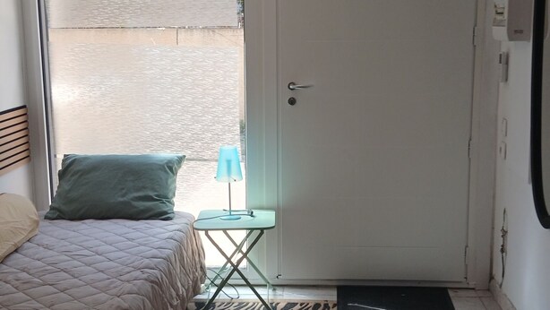 A bedroom with a bed, a small table, a door, and a view of the outside. - Studio 16 mcarré kicnetteet frigo compartiment congelateur micro ondes (Perpignan,)