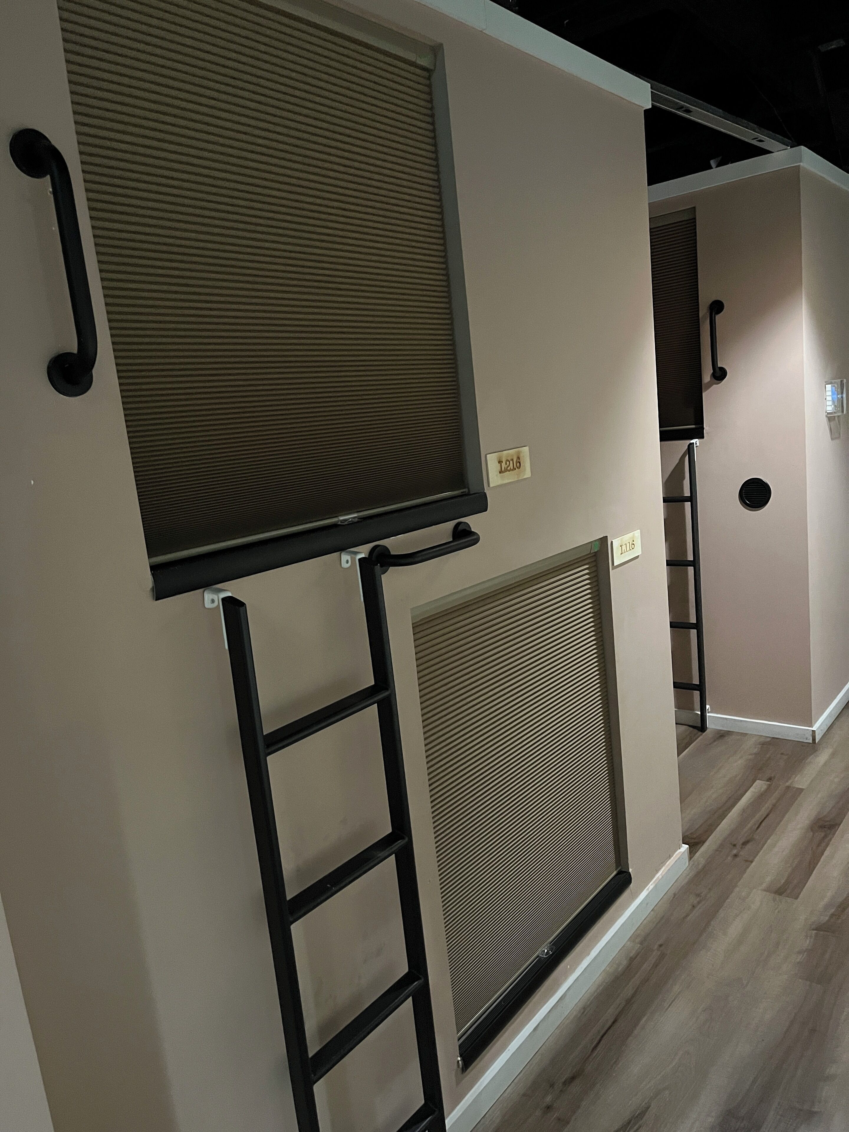 A room with beige walls, a wooden floor, and two large windows with blinds.