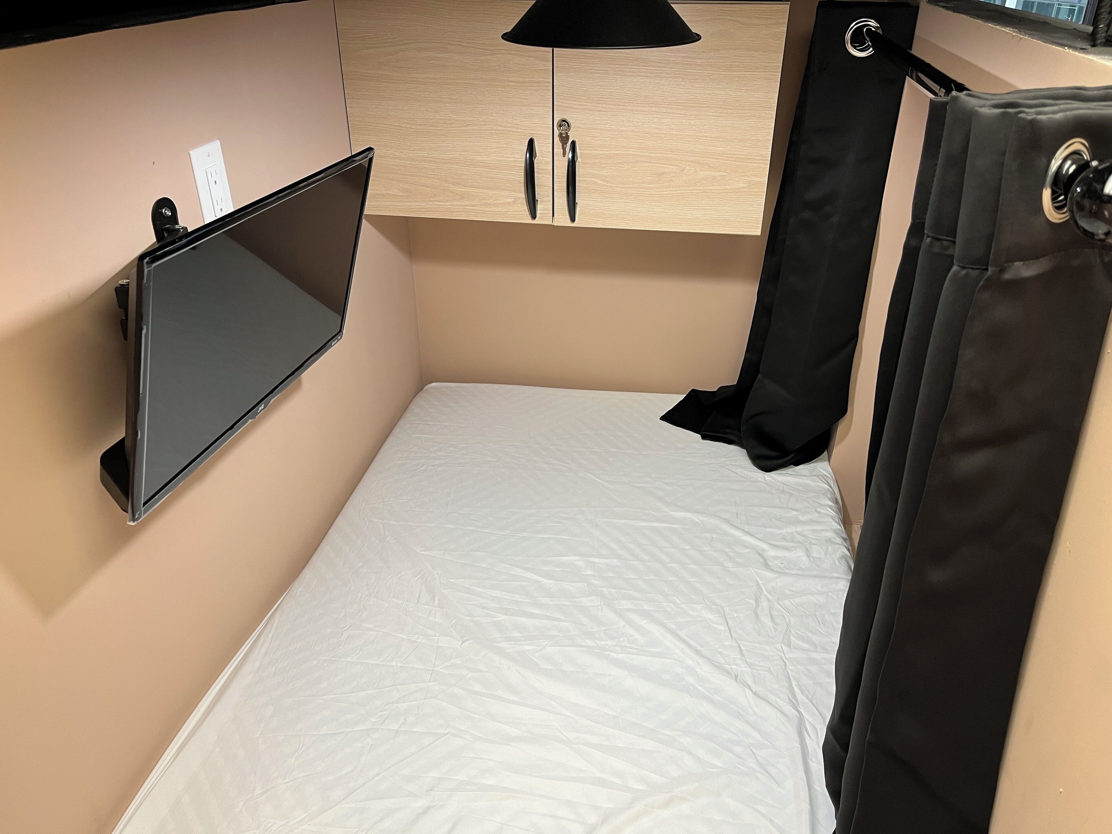 A compact room with a bed, a mounted TV, a wooden cabinet, and a black curtain.