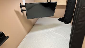 Una habitación pequeña con una cama, un televisor montado, un estante y un armario de madera.