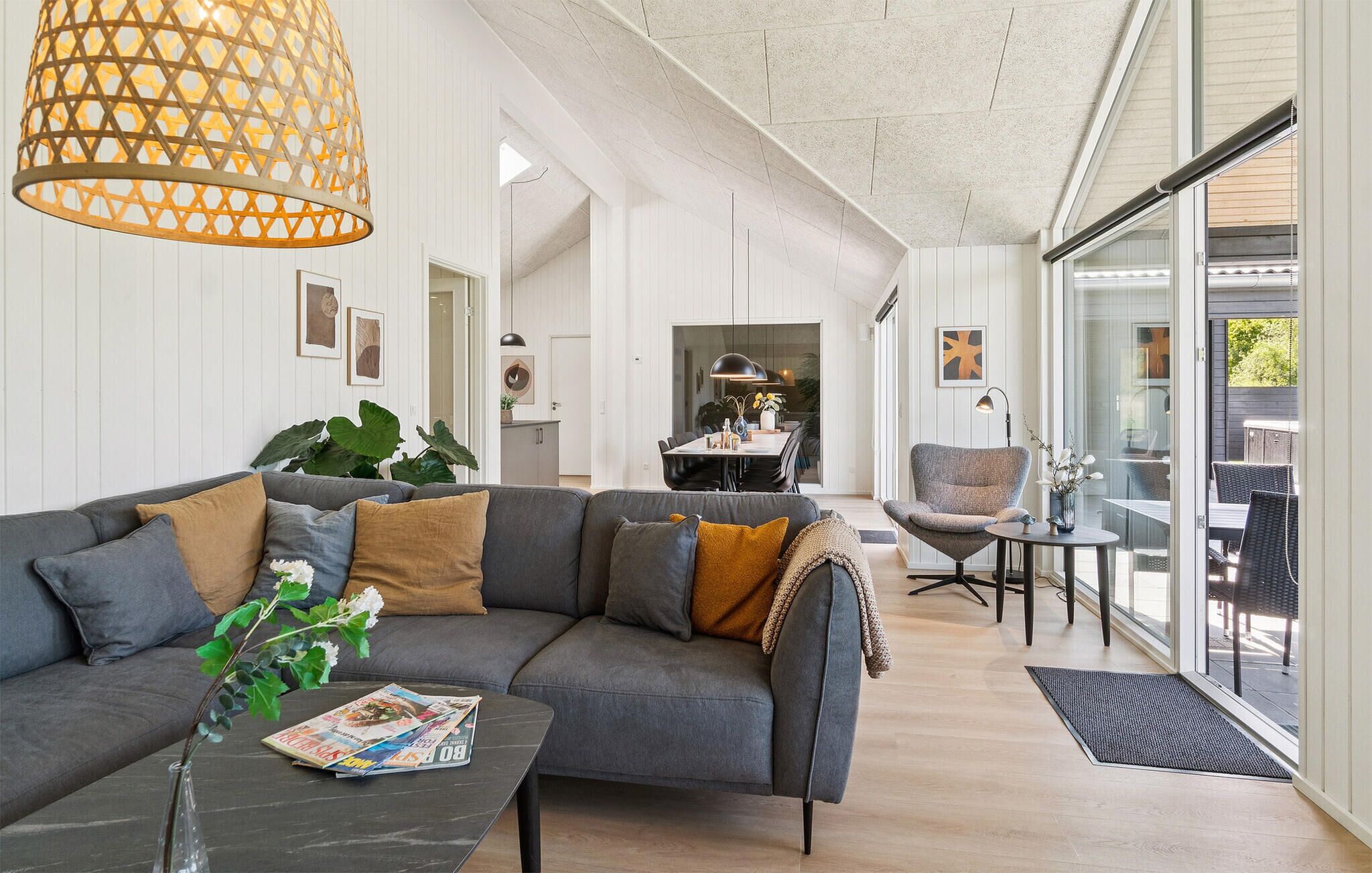 Ett modernt vardagsrum med en grå hörnsoffa, ett soffbord med tidningar, en hängande lampa och utsikt över matplatsen genom glasdörrar.