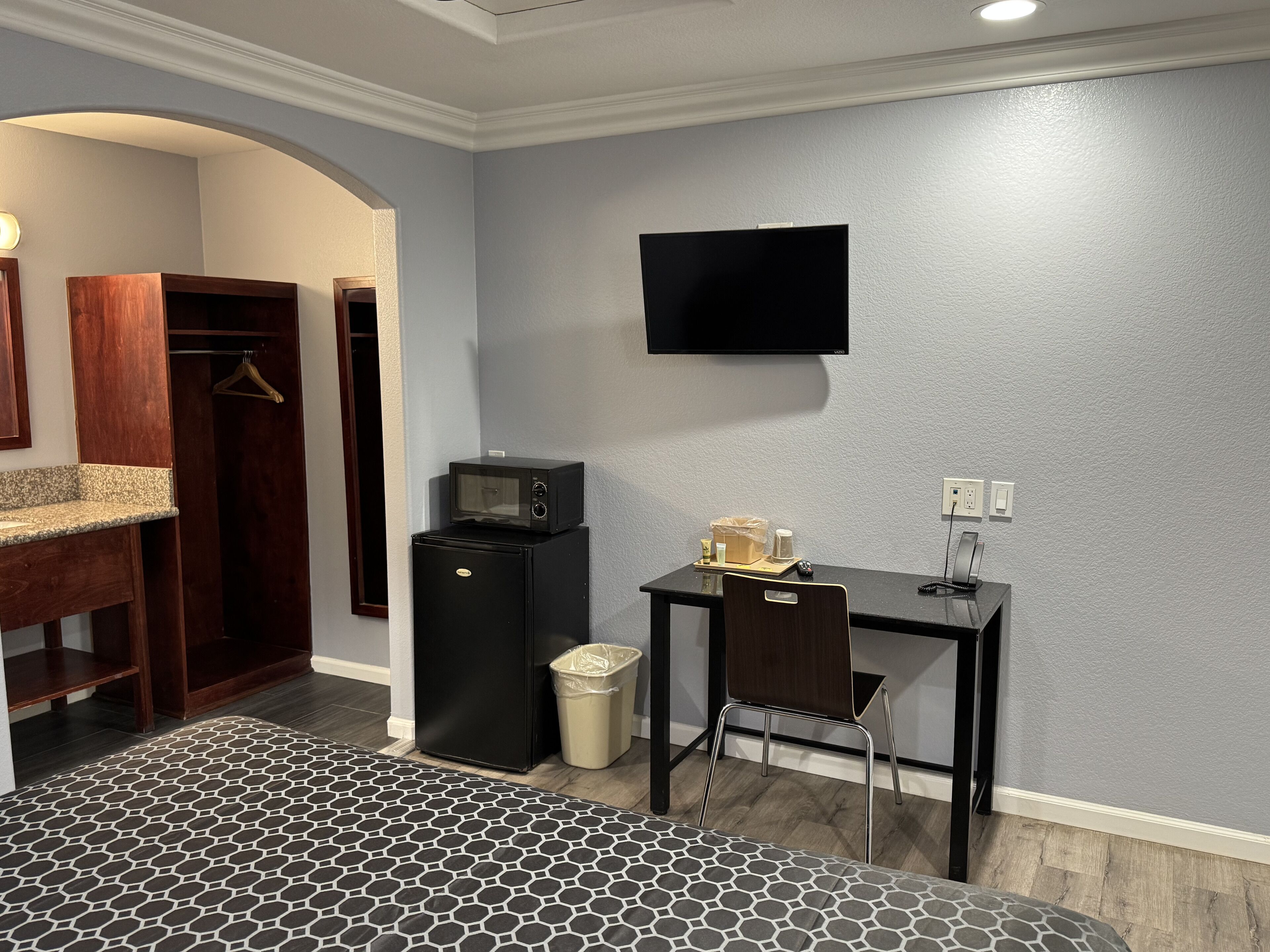 A compact hotel room with a kitchenette, a bed with a patterned bedspread, a TV mounted on the wall, and a small trash bin.