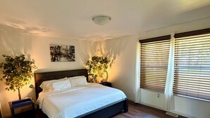 Ein Schlafzimmer mit einem Bett, zwei Nachttischen, einer Wandleuchte, einem Fenster mit Jalousien und einer Topfpflanze.