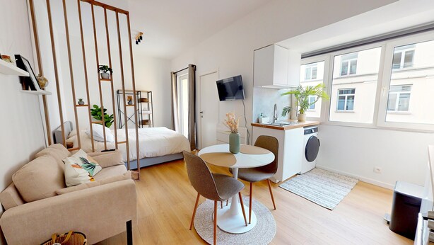 A modern, well-lit room with a bed, a sofa, a dining table with chairs, a small kitchen area, and a large window providing a view of buildings outside. - Studio cosy centre avec balcon (Brussels)