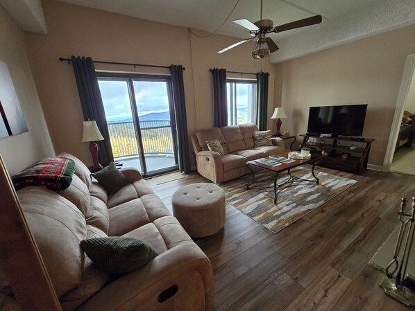 A spacious living room with a sofa, ottoman, and a coffee table. There is a television, a lamp, and a ceiling fan.