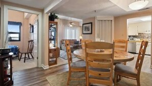 A dining area with a round wooden table and chairs, a kitchen in the background, and a view of the outdoors through a door. - Charming Sky Blue 2-Bedroom Condo in Sugar Mountain with Resort Amenities (Sugar Mountain)
