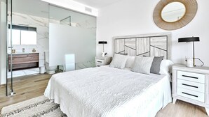 Quarto moderno com uma cama grande, mesa de cabeceira, espelho e banheiro com porta de vidro.