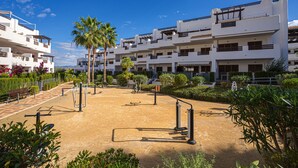 Un bâtiment moderne doté de nombreux balcons, d’une cour avec des équipements de musculation et de palmiers.