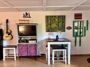 Une pièce comprenant une télévision, une guitare, un meuble, une table avec des chaises et un tableau.