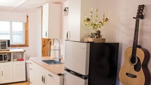 Een kleine keuken met witte kasten, een zwarte koelkast, een magnetron en een gitaar die tegen de muur staat.