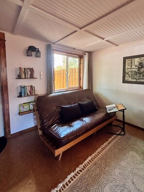 A room with a brown leather sofa, a small table, a bookshelf, a window with a view of a wooden fence, and a framed picture on the wall.