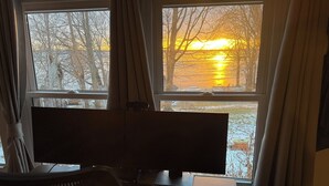 A room with a desk, a chair, and a computer monitor. There is a window with curtains, and a view of trees and a sunset. - Lakefront Retreat w/ Kayaks, Firepit & Home Office.  Beautiful Sunsets! (Constantia)
