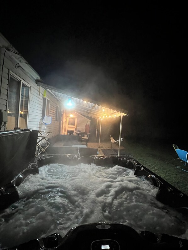 Un jacuzzi con agua corriendo, un porche cubierto con luces colgantes y una casa al fondo.