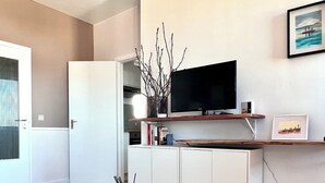 A modern living room with a glass coffee table, a flat-screen TV mounted on a wooden shelf, and a white cabinet in the background. - Appartement Proche de l'Otan (Brussels)
