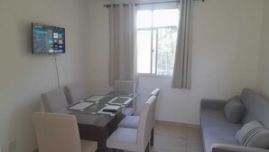 Un área de comedor con una mesa y sillas, un televisor montado en la pared y un ventanal con cortinas.