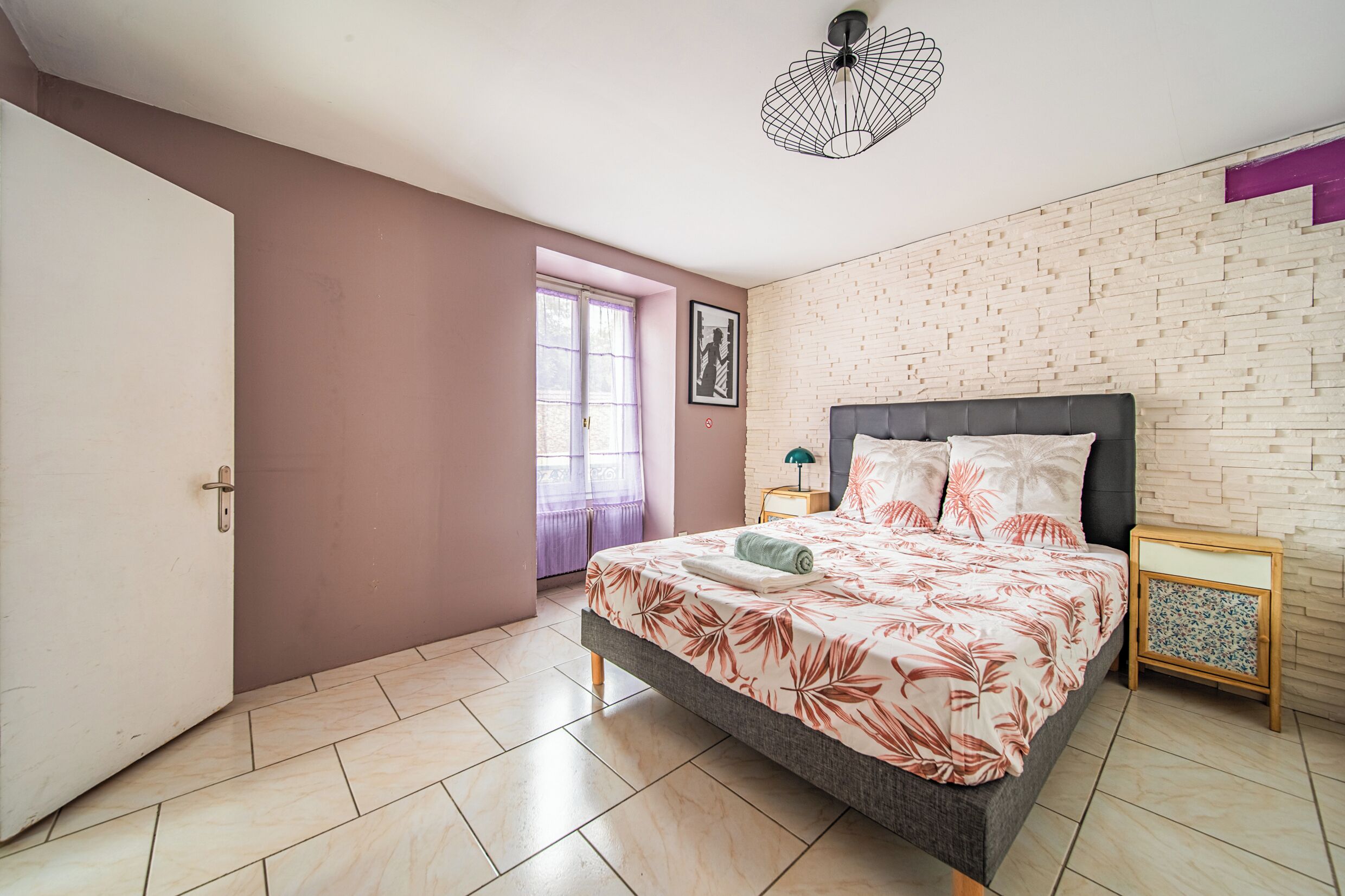 A bedroom with a bed, a nightstand, a window with curtains, a door, and a wall with a brick pattern.
