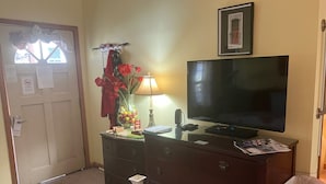 Ein Zimmer mit Fernseher auf einem Schrank, einem Schminktisch, einer Lampe, einer Vase mit Blumen und einem Mülleimer.