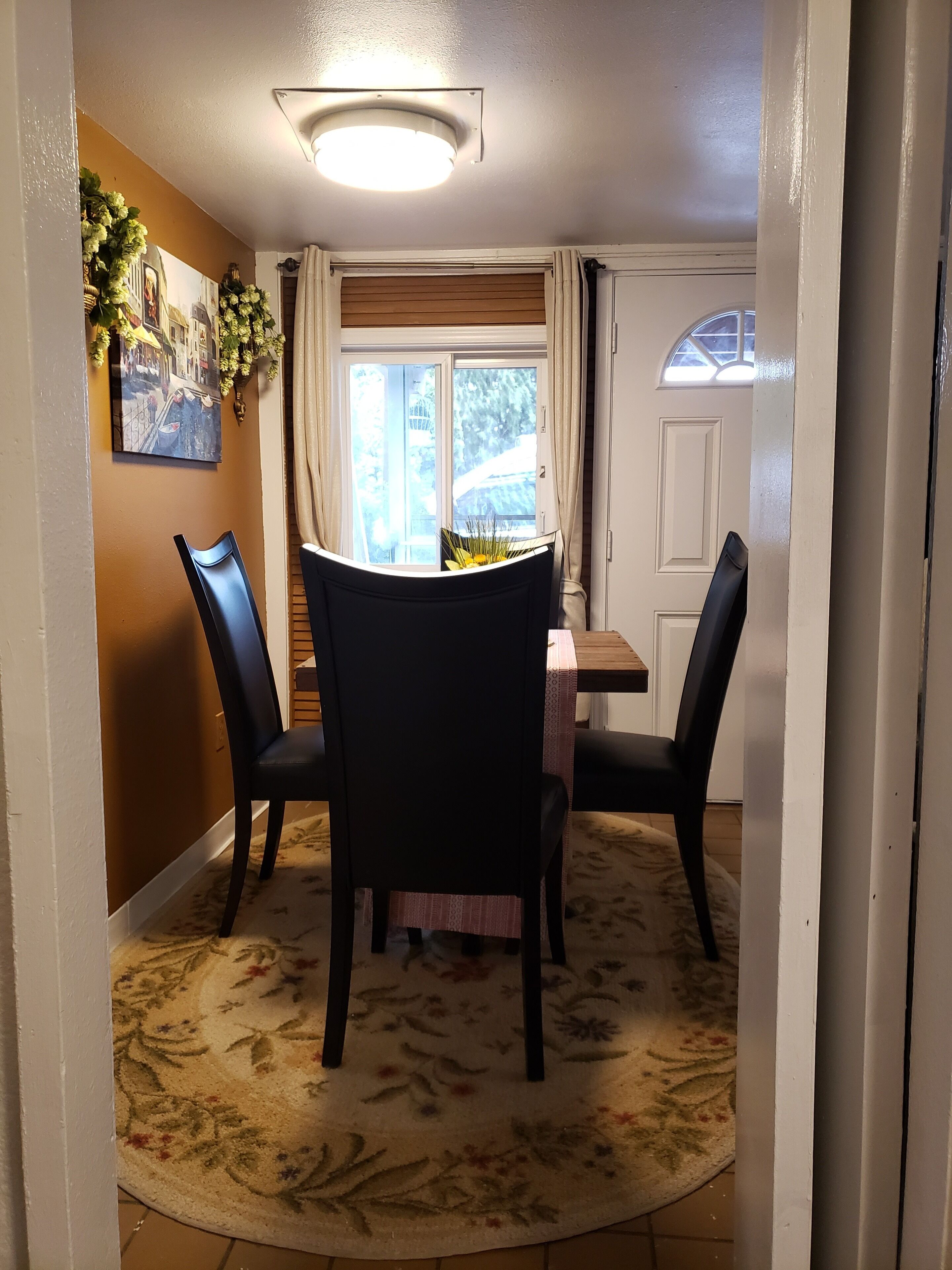 A dining area with a round table, four chairs, a floral rug, a ceiling light, a window with curtains, and a door.