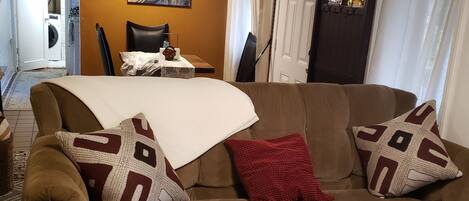 A living room with a brown sofa, patterned cushions, a white throw blanket, a small table with a vase of flowers, and a painting on the wall.