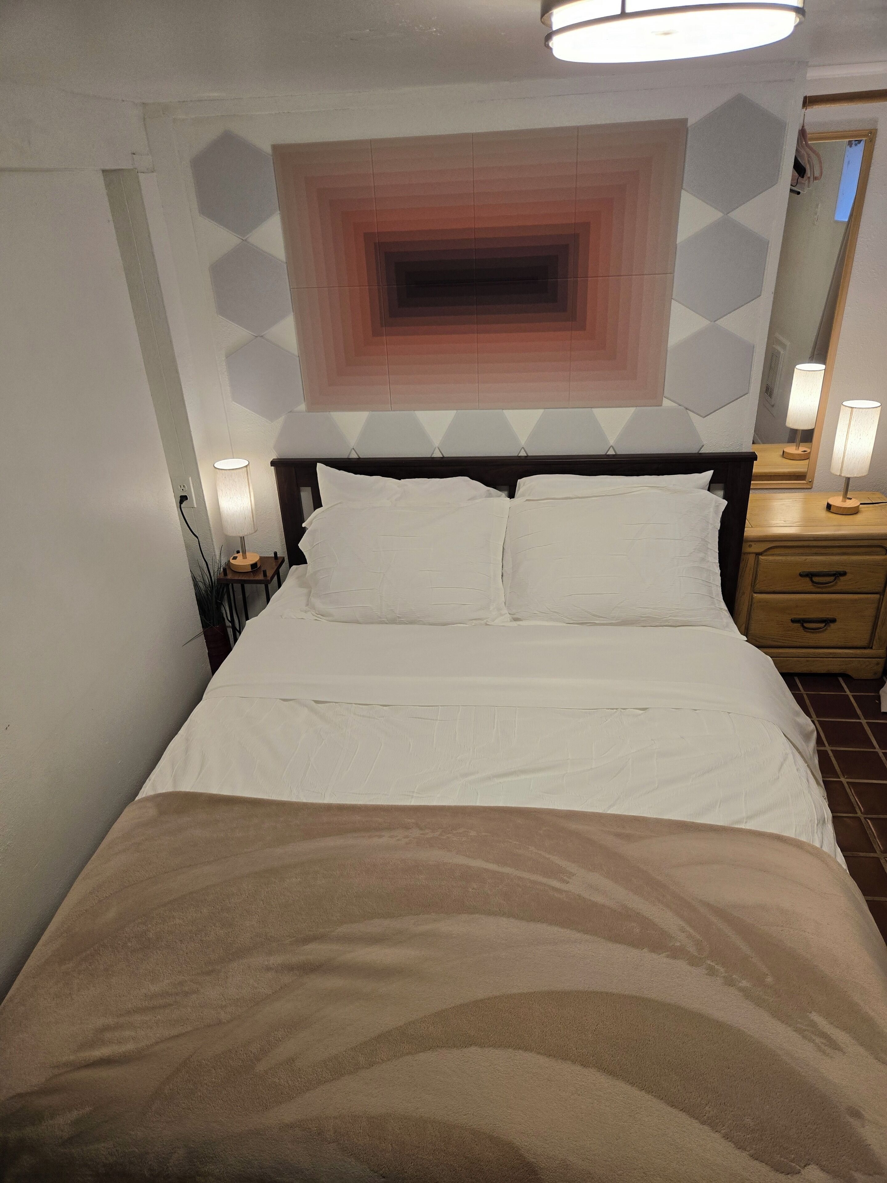 A hotel room with a large bed, a nightstand with a lamp, a dresser, and a geometric-patterned wall above the bed.