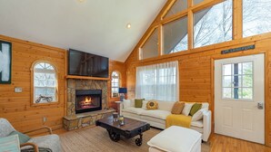 A cozy cabin interior with a stone fireplace, wooden walls, a sofa, and a television. - Deer Haven Cabin, Stunning Mtn Views, Hot Tub+Fire Table+Grill, Game Room, WiFi! (Burnsville)