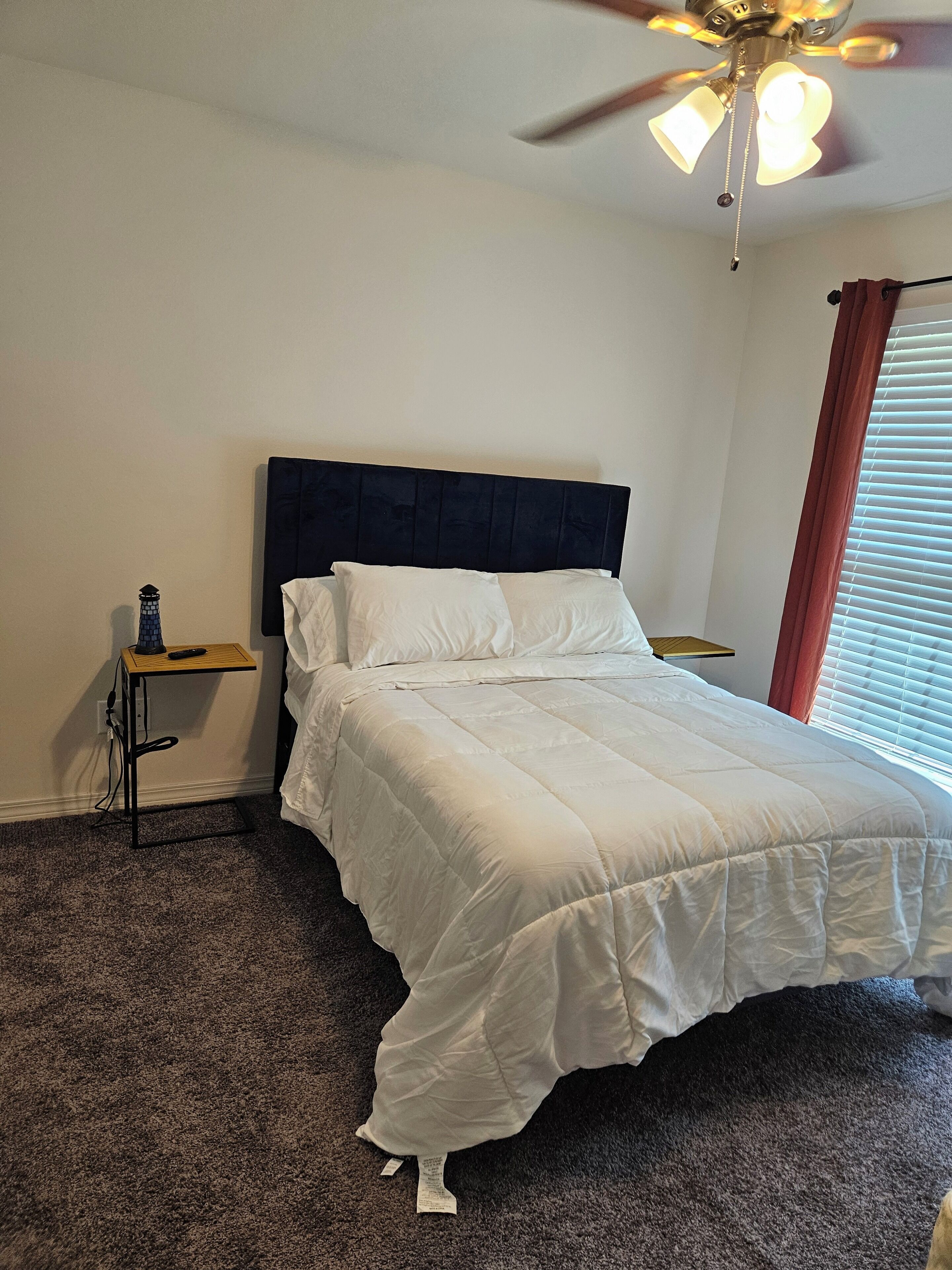 Una cama bien hecha en una habitación con ventilador de techo, una mesita de noche pequeña con lámpara y una ventana con persianas.