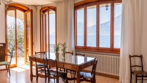 A dining area with a wooden table and chairs, large windows with curtains, and a view of the sea and mountains. - VILLA ESMERALDA BETH'S HOUSE (Brenzone sul Garda)