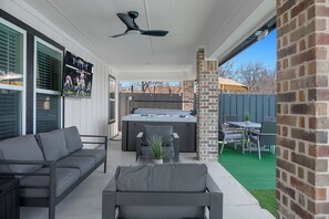 A covered patio area with a hot tub, outdoor seating, and a TV screen displaying a sports event.