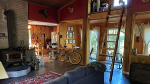 A cozy cabin interior with a sofa, a wooden stove, a bicycle, and a ladder leading to a loft. - Dragon’s Nest on River. Quiet Get-away (Tweed)
