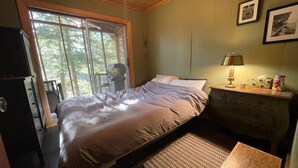 A bedroom with a bed, a nightstand, a lamp, and a window with a view of trees. - Dragon’s Nest on River. Quiet Get-away (Tweed)