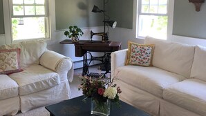 Una sala de estar con dos sofás blancos, una mesa de centro oscura con un florero, una máquina de coser y un ventilador de techo.