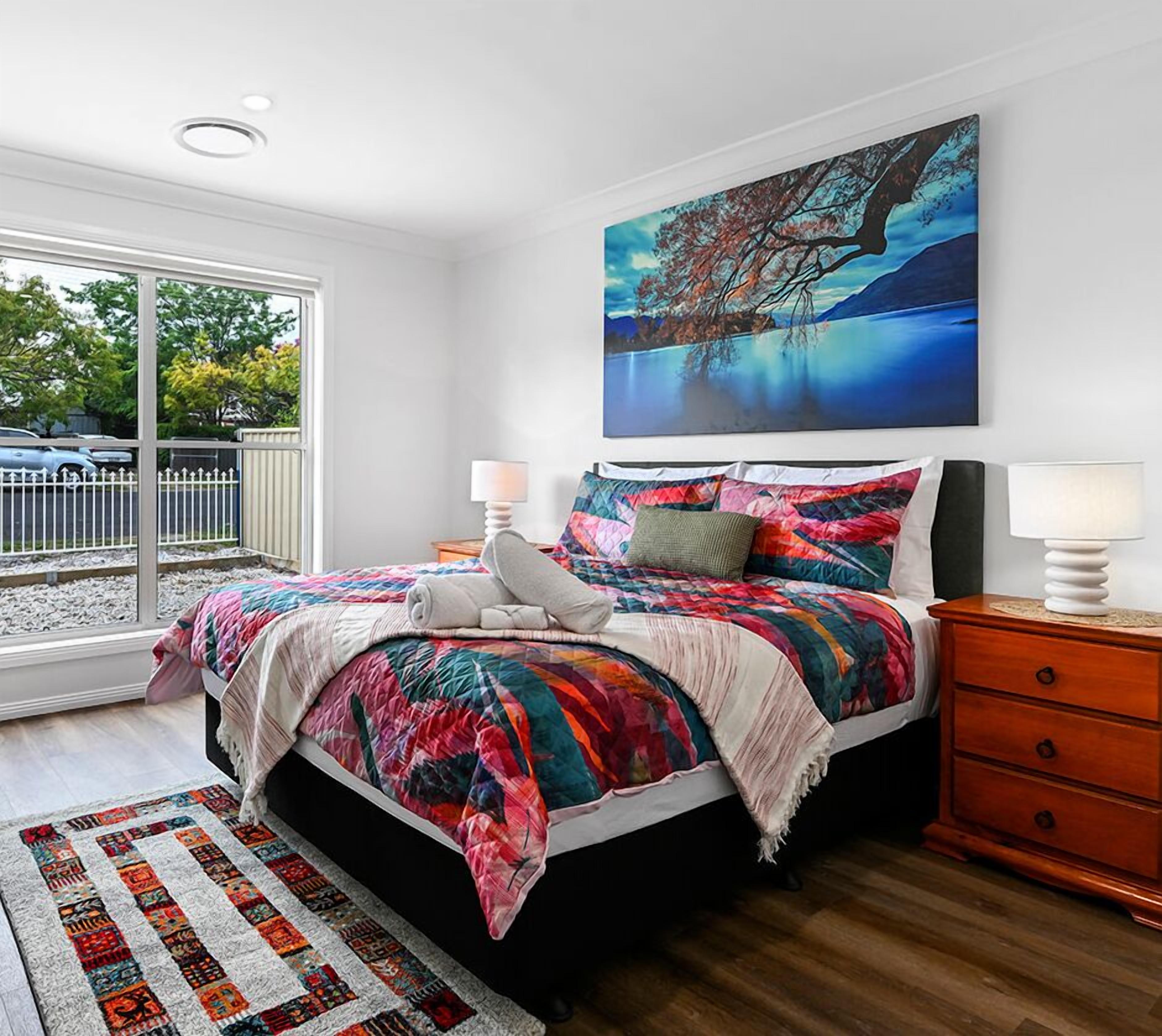 A bedroom with a bed, a nightstand, a lamp, a painting, and a view of the outside.