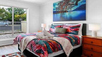 A bedroom with a bed, a nightstand, a lamp, a painting, and a view of the outside.