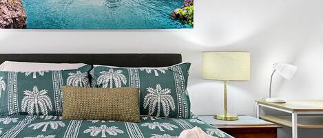 A hotel room with a bed featuring a tropical palm tree pattern, a view of a tropical beach, and a bedside table with a lamp.