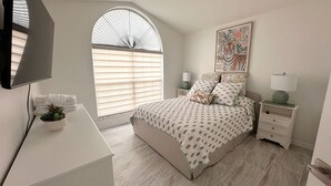 Ein Schlafzimmer mit Bett, Schreibtisch, Fernseher, einem Fenster mit Jalousien, einem Nachttisch, einer Lampe und einem gerahmten Bild an der Wand.