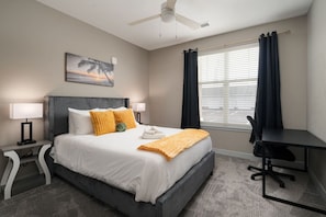 Une chambre d’hôtel avec un lit, un bureau, une chaise, un ventilateur de plafond, une fenêtre avec des rideaux et un tableau au mur.