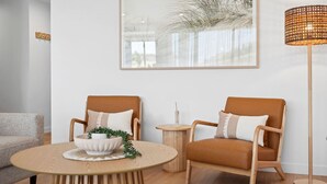 Una sala de estar moderna con dos sillones de cuero marrón, una mesa redonda de madera con un cuenco blanco y plantas verdes, una pequeña mesita auxiliar de madera, y una lámpara de pie alta.