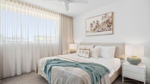 Un dormitorio con una cama grande, dos mesitas de noche con lámparas, un ventilador de techo y un cuadro con motivos playeros en la pared.