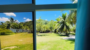 A view through a window of a grassy area with palm trees and a small structure. - FARE HIRO BUNGALOW 1 SEASIDE (Moorea-Maiao)