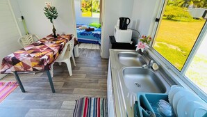 A compact room with a kitchenette, a dining area with a table and chairs, a bed with a blue and white patterned bedspread, and a window with a view of a grassy area. - FARE HIRO BUNGALOW 1 SEASIDE (Moorea-Maiao)