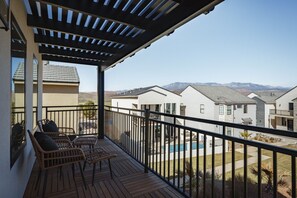 A balcony with a wooden deck, wicker chairs, and a view of neighboring houses and mountains.