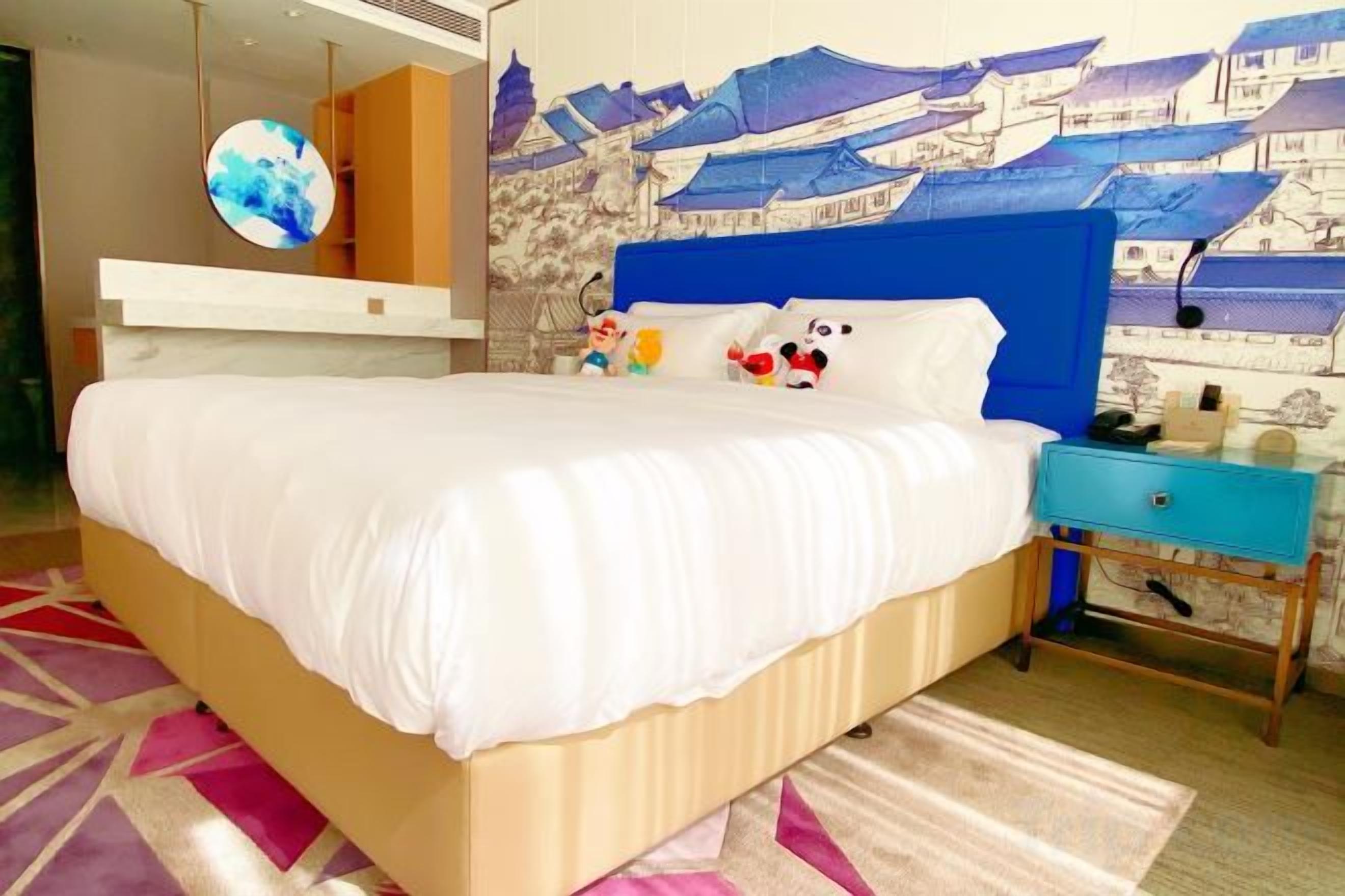 A hotel room with a bed, a nightstand, a wall with a mural, and a round mirror.