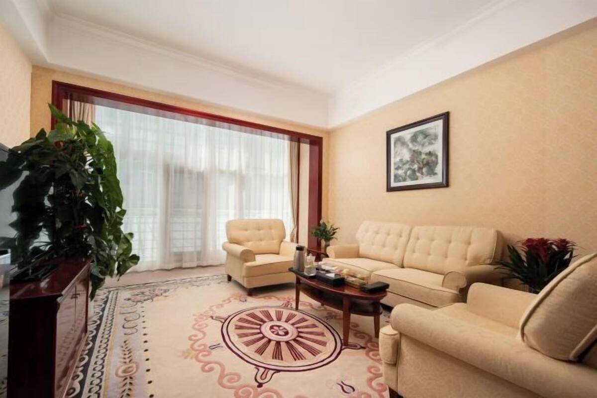 A living room with a beige sofa, a chair, a coffee table, a large potted plant, and a framed picture on the wall.