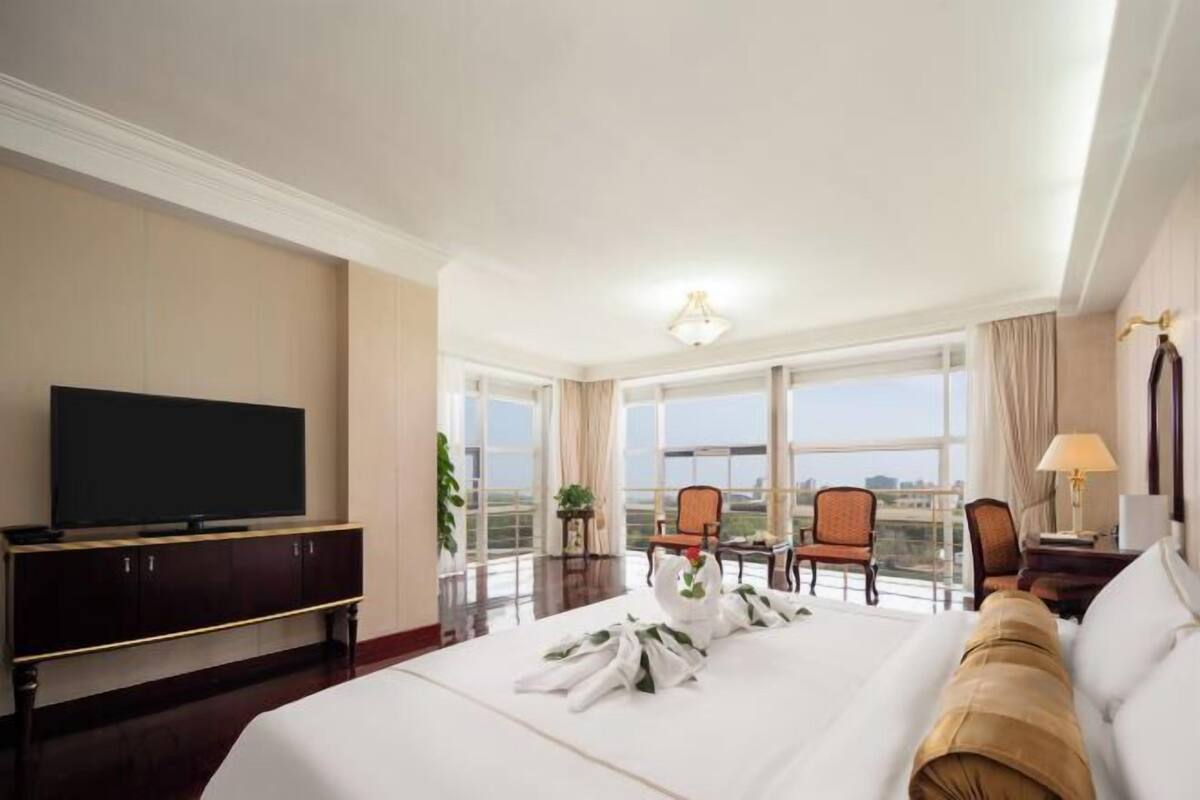 A hotel room with a large bed, a television on a cabinet, a balcony with chairs, and a view of the city.