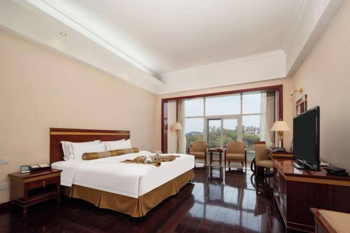 A hotel room with a large bed, a desk with a TV, a chair, and a window with a view.
