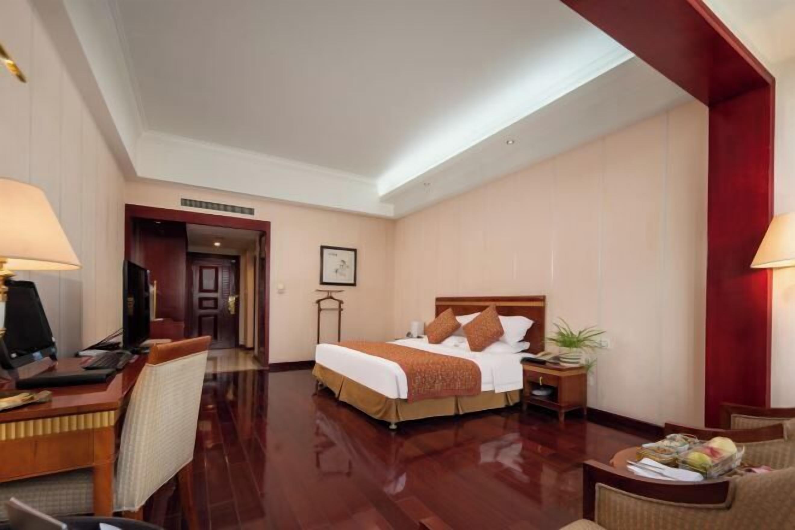 A hotel room with a large bed, a desk with a computer, a chair, a small table with a lamp, and a plant.