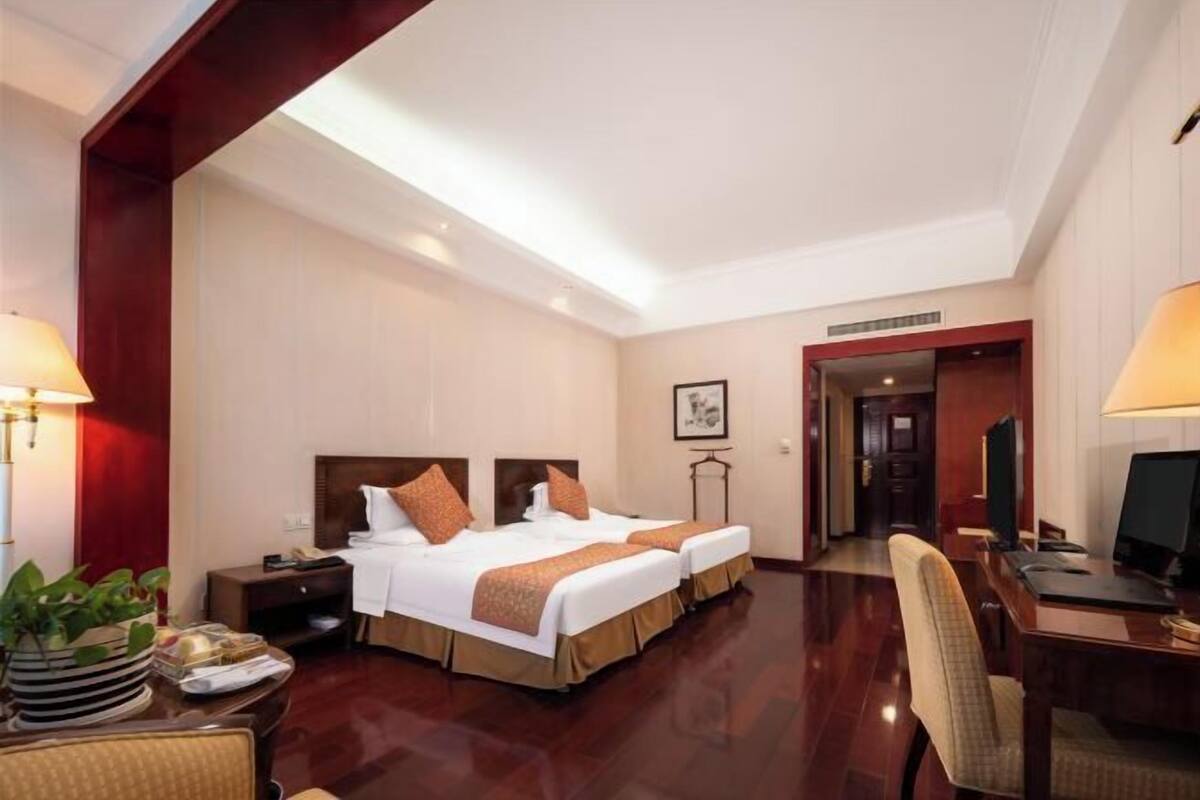 A hotel room with two beds, a desk, a chair, a TV, and a lamp.