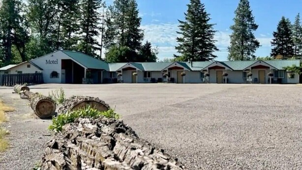 Un motel composé de plusieurs bâtiments, un parking en gravier et entouré de grands arbres.