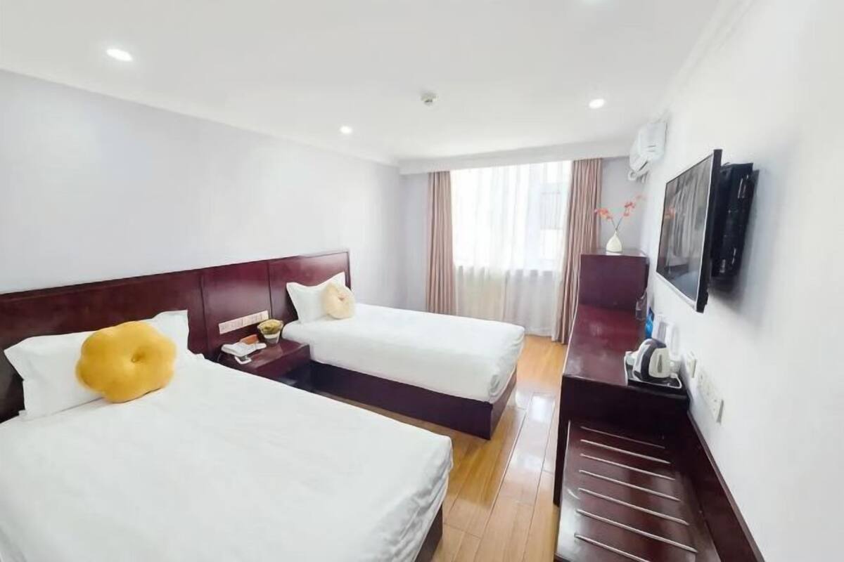 Une chambre d’hôtel avec deux lits, une tête de lit en bois, un téléviseur à écran plat, un bureau et une fenêtre avec des rideaux.